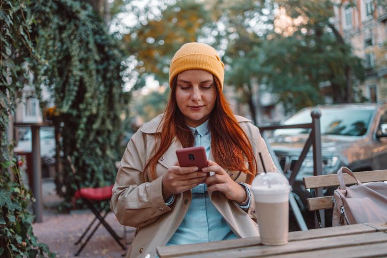 Vrouw zit met koffie op terras en kijkt op telefoon